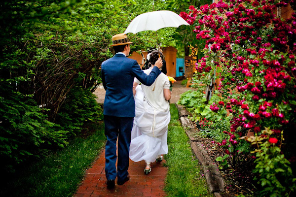 cute photo of the bride and groom running through a garden to get out of the rain while the groom holds a white umbrella over the bride - groom is wearing blue suit with a straw derby hat - photo by New Mexico based wedding photographers Twin Lens